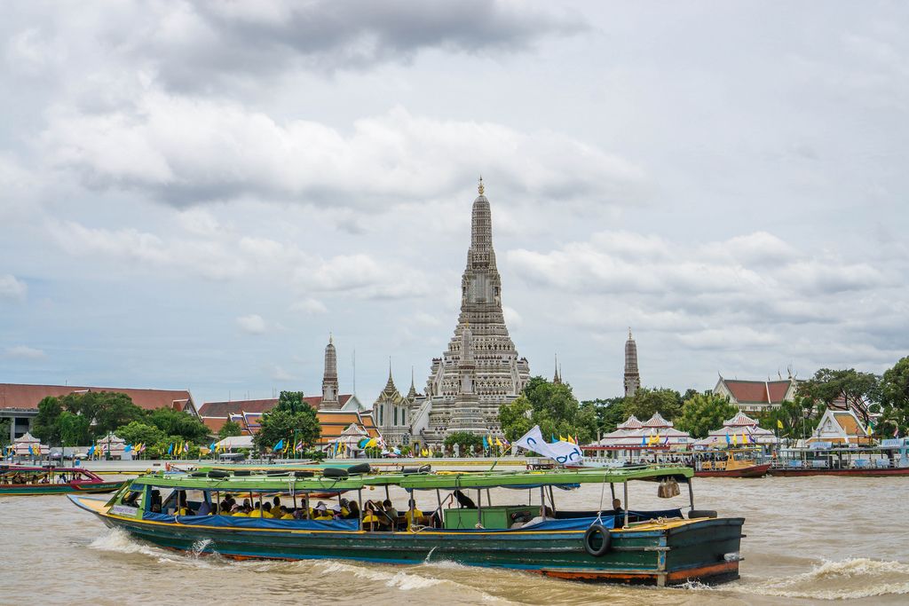 Boat with Tourists driving past Wat Arun Temple on Chao Phraya River in Bangkok