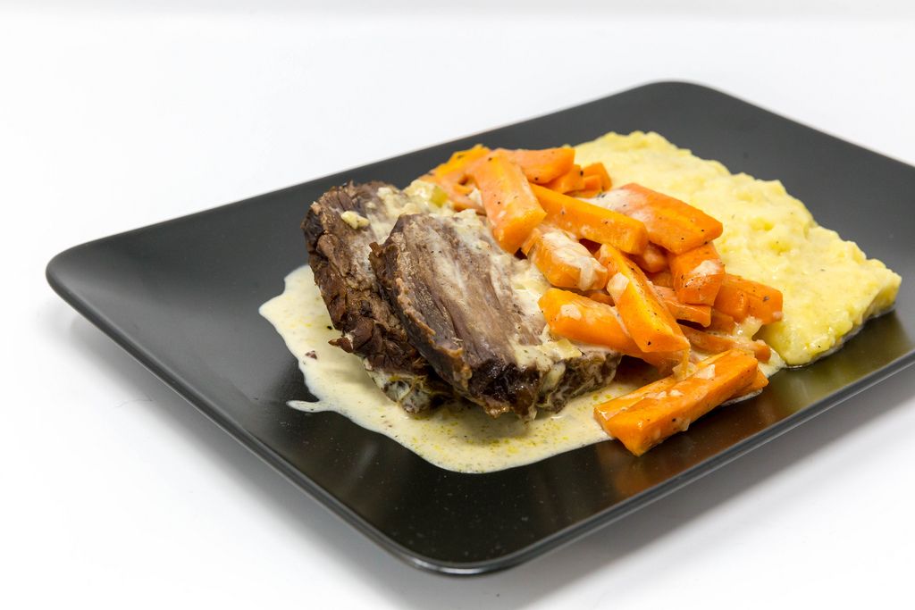 Boiled beef with steamed carrots, mashed potato and sauce on a black plate