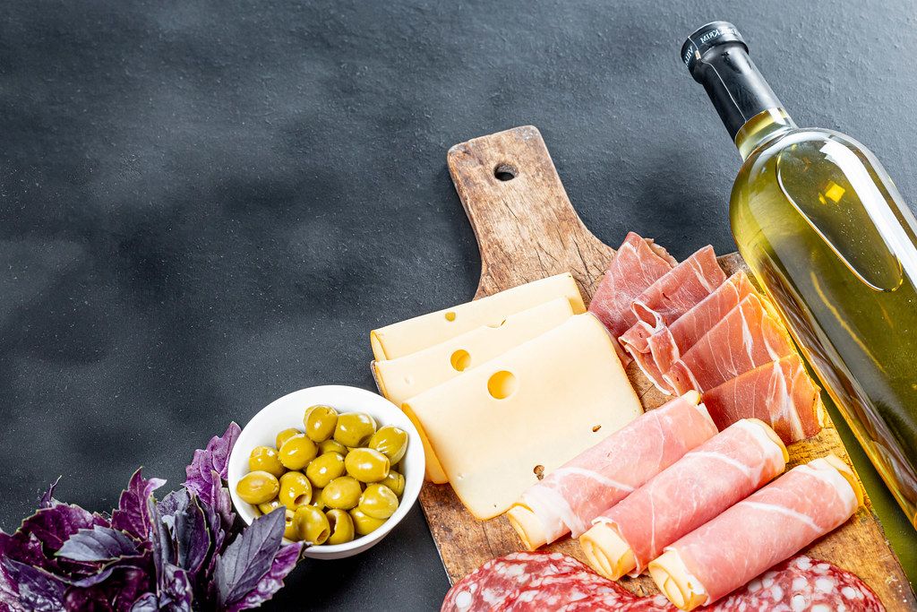 Bottle of white wine, fresh Basil, cold cuts, cheese with green pickled olives on black background (Flip 2019)