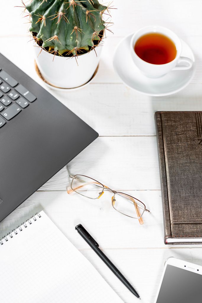 Büroarbeitsplatz mit Laptop, Notizblock, Kalenderbuch, Brille, Mobiltelefon, einer Tasse Tee und Kaktus