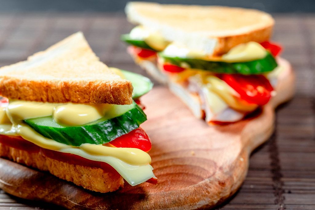 Close Up Food Photo of halved Ham Sandwiches with Melted Cheese, Cucumber and Tomato on a wooden Board