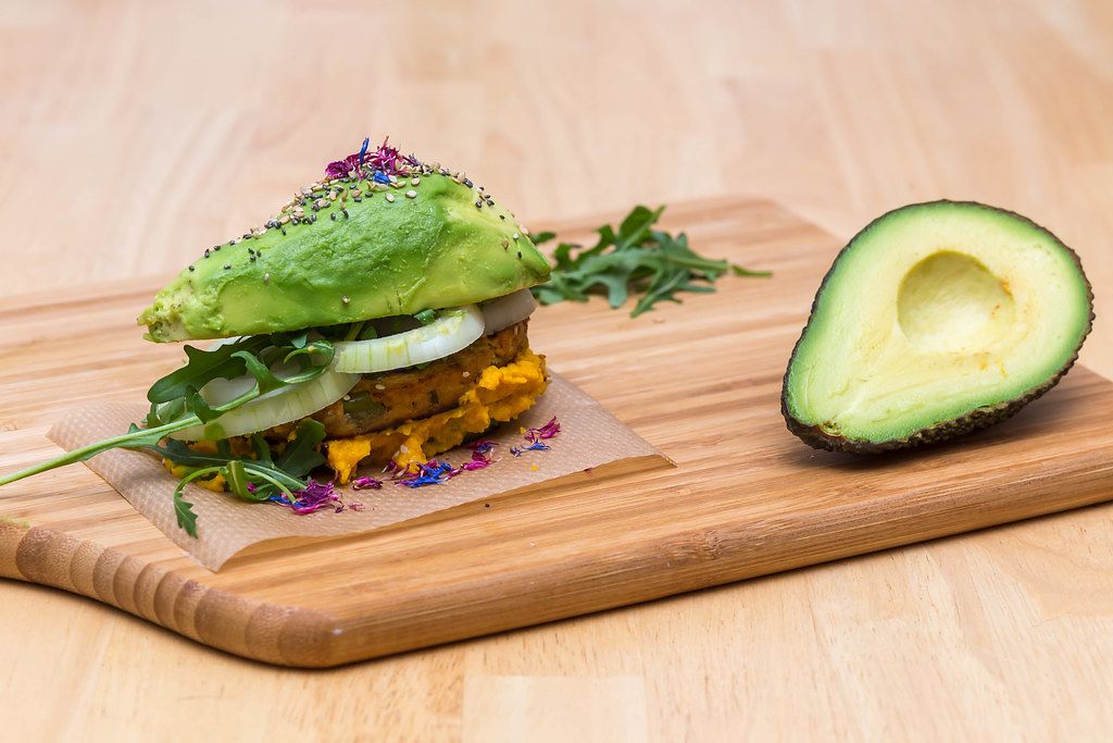 Close Up Food Photo of Home Made Vegetarian Avocado Burger with Hummus and Fresh Onion next to Halved Avocado on Wooden Cutting Board