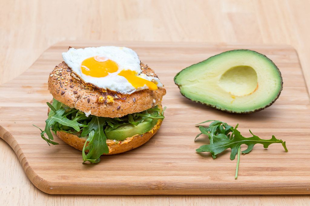 Close Up Food Photo of Vegetarian Avocado Burger with Arugula and Sunny Side Up Egg next to Halved Avocado on Wooden Cutting Board