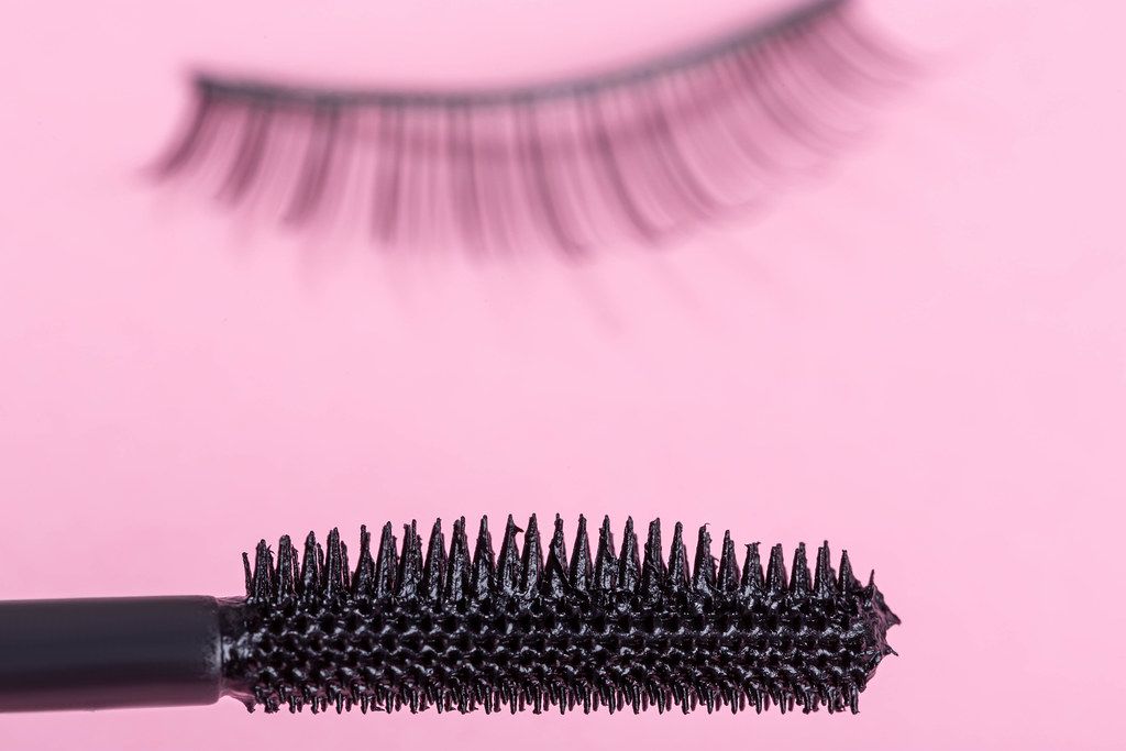 Close up of a mascara brush on a pink background with false lashes behind