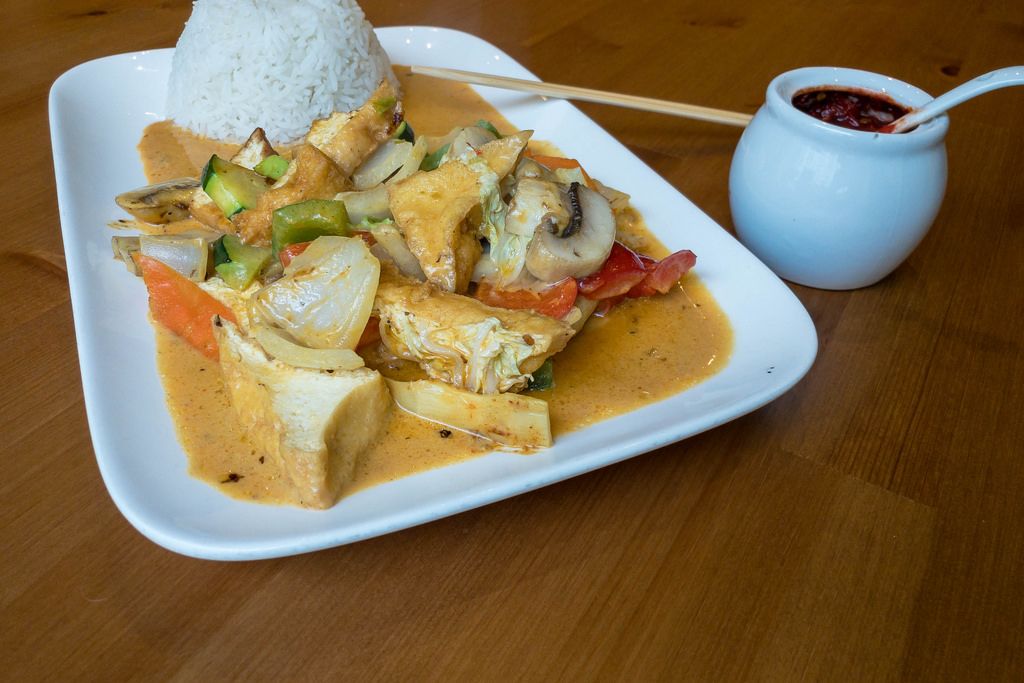Curry-Pfanne mit Tofu, Pilzen, Karotten Zucchini und frischen Paprika mit Reis aus einem Teller mit Essstäbchen und Chilipaste