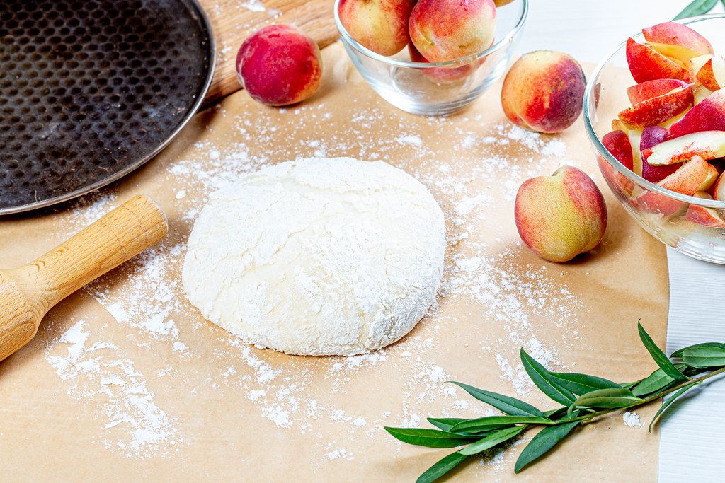 Dough, flour and fresh peaches with rolling pin and baking sheet on the table (Flip 2019)
