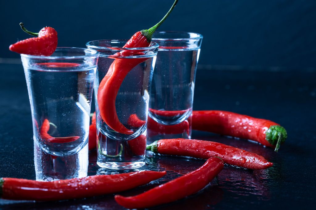 Drei kleine Gläser gefüllt mit Wodka mit scharfen roten Chilis vor schwarzem Hintergrund