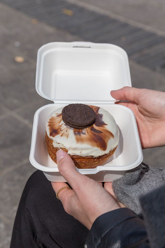 Ein Kronut-Donut aus Blätterteig, mit weißem Schokoguss in einer weißen Burgerbox, aus dem Feinkostgeschäft 