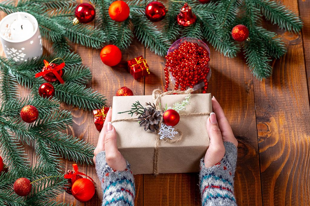 Eine Frau mit Stulpen an den Händen hält ein Weihnachtsgeschenk, umgeben von Tannenzweigen und roten Weihnachtsbaumkugeln