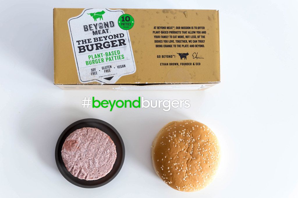 Flatlay of vegan burger patties in the Beyond Meat Burger 10pcs Box, with frozen soy-free and gluten-free patties, next to a burger roll, on white surface