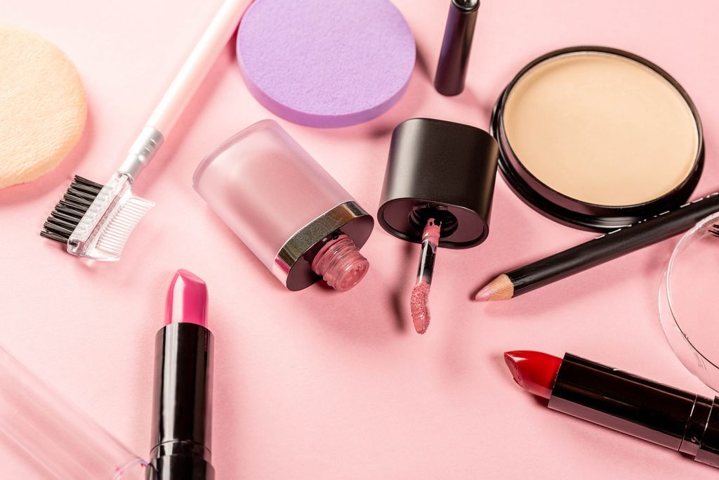 Frauenkosmetik wie Puder, Lippenstift, Lipgloss, Lipliner und Brauenbürste vor rosa Hintergrund