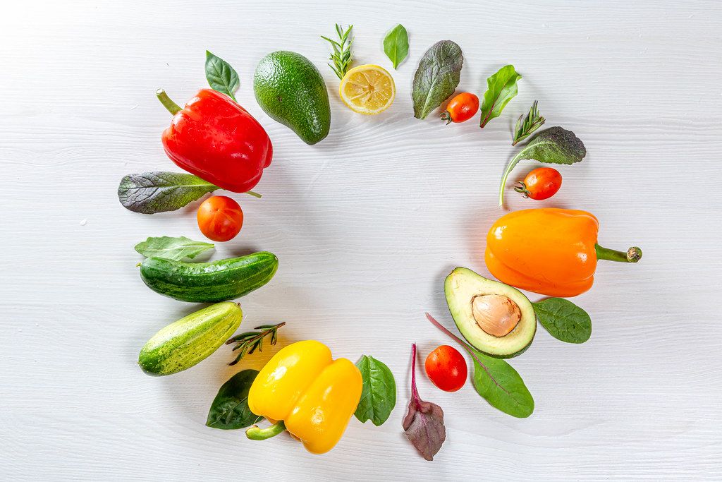 Fresh vegetables and greens are laid out in a circle on a white wooden background. Top view