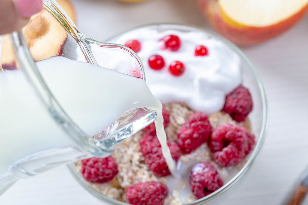 From a glass jug pour milk into oatmeal with fruit