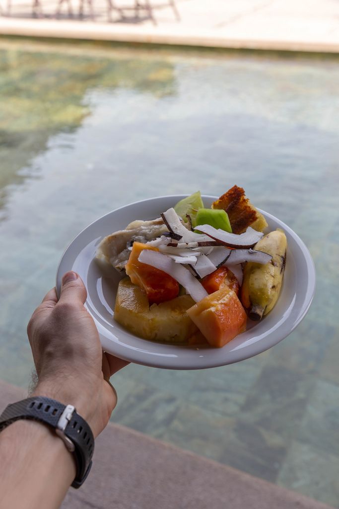 Fruit salad with banana, papaya, pineapple and coconut on white plate hold above the pool of the Constance Ephelia Resort in Mahé, Seychelles