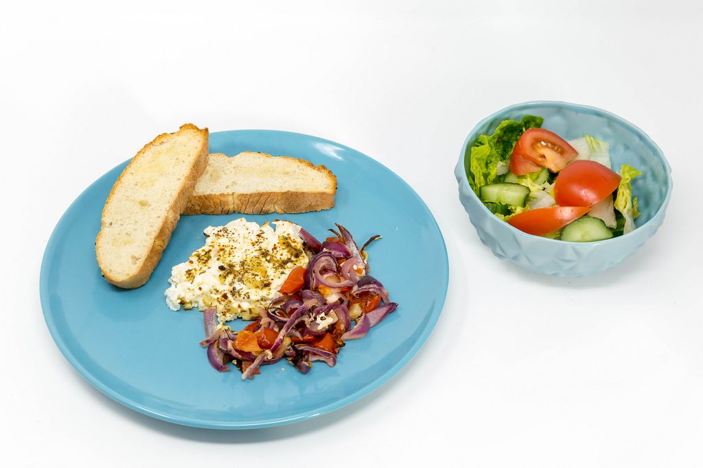 Gebackener Hirtenkäse an Tomatengemüse dazu Knoblauchfladenbrot auf einem blauen Teller und ein frischer Salat auf weißem Hintergrund