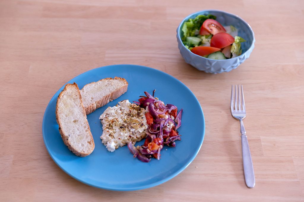 Gebackener Hirtenkäse an Tomatengemüse dazu Knoblauchfladenbrot auf einem blauen Teller und ein frischer Salat