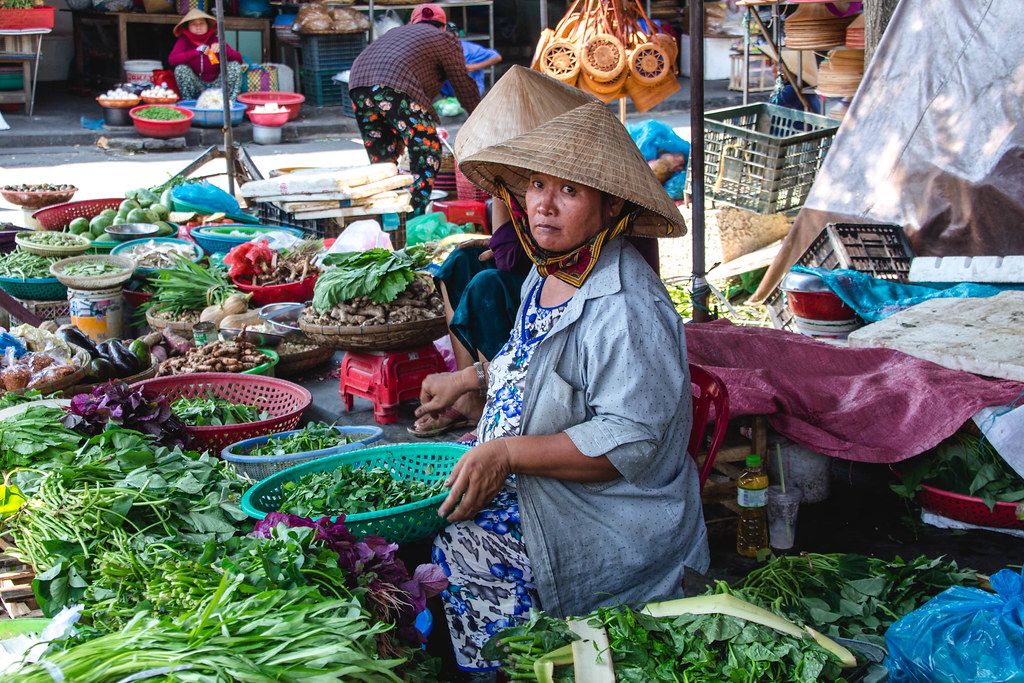 Gemüse-Verkäuferin mit geflochtenem Kegelhut auf einem Markt in Hoi Ann
