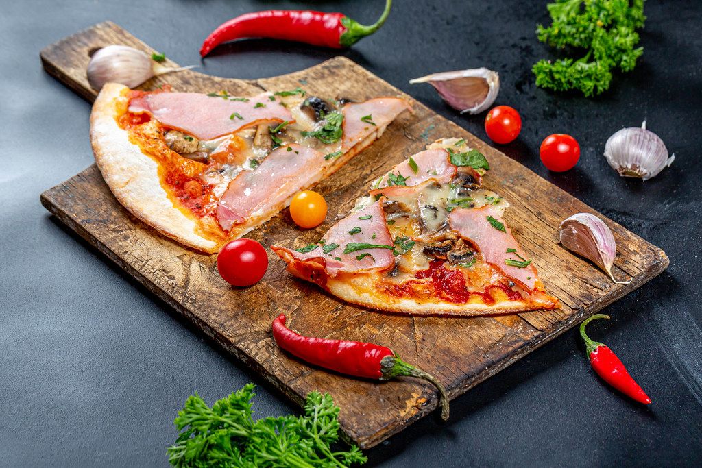 Ham pizza slices on an old wooden kitchen Board with tomatoes, chili and parsley