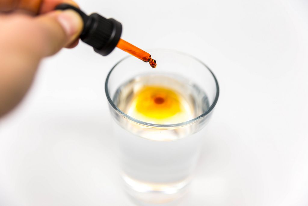 Hand drips one single drop of Braineffect Recover Cannabidiol (CBD) oil in a glas and it turns water orange in front of white background