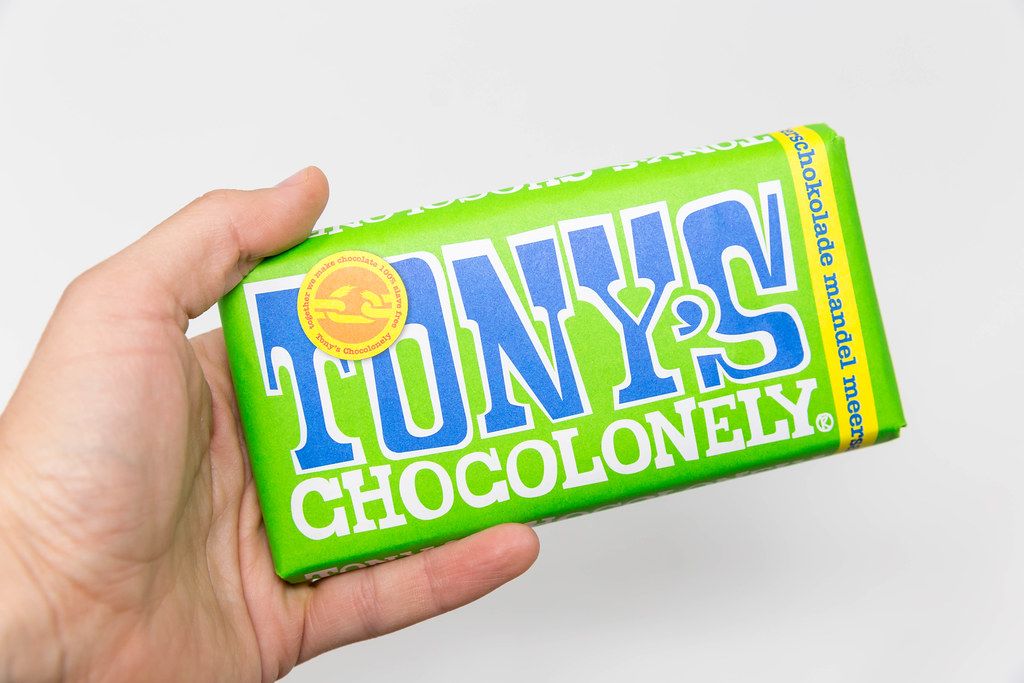 Hand hält eine 100% sklavenfreie Tony's Chocolonely Schokoladentafel mit Mandeln und Meersalz. Grüne Packung vor weißem Hintergrund