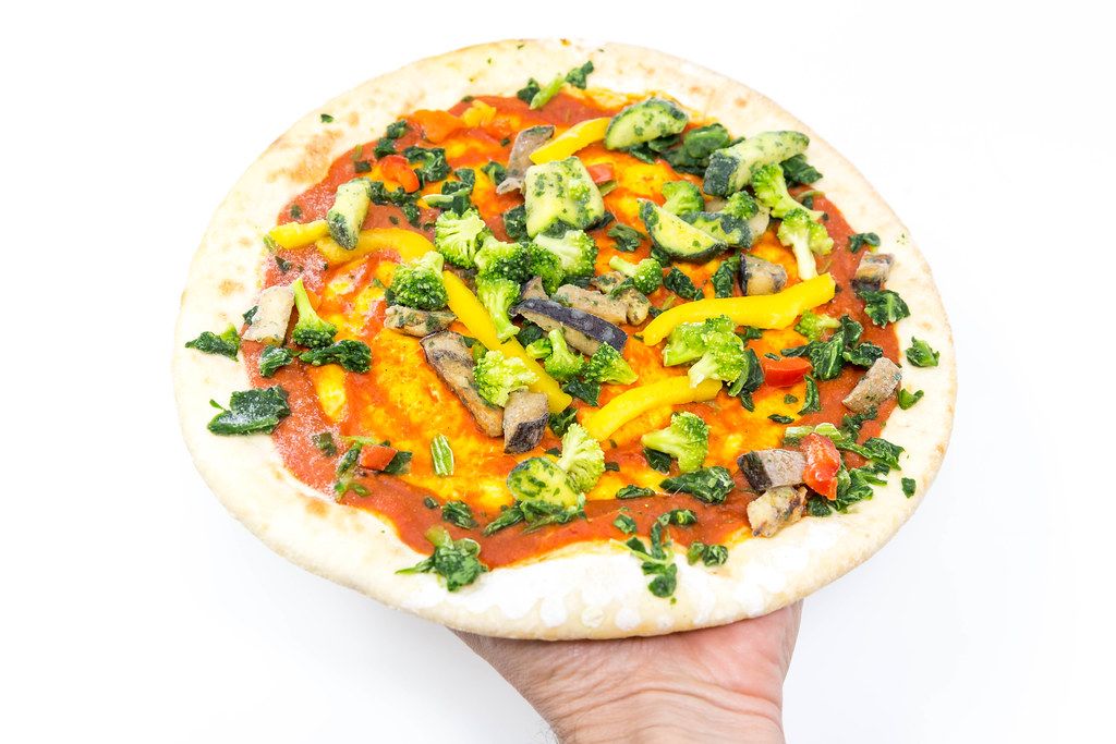 Hand holds a frozen vegan pizza with fresh broccoli and grilled vegetables on a white background