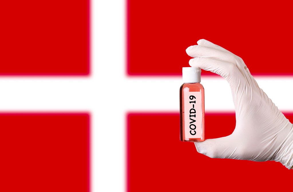 Hand in protective gloves holding COVID-19 test tube in front of flag of Denmark