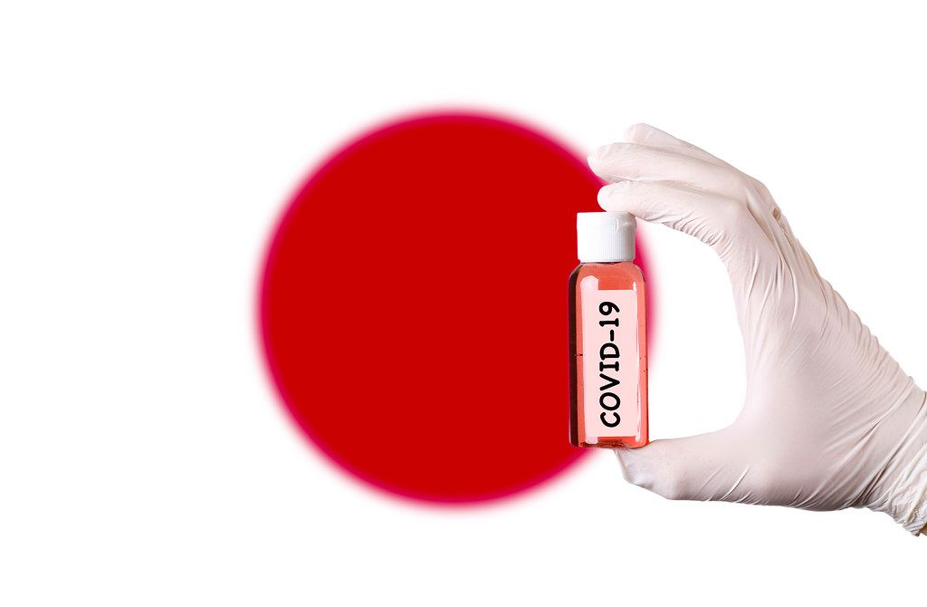 Hand in protective gloves holding COVID-19 test tube in front of flag of Japan