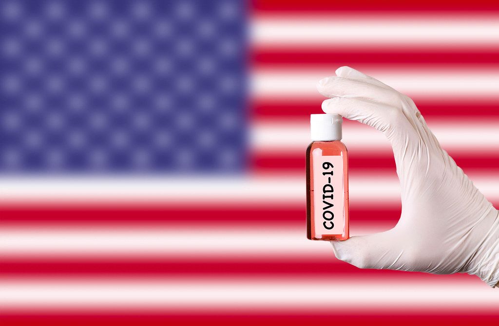 Hand in protective gloves holding COVID-19 test tube in front of flag of USA