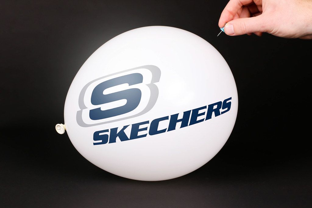 Hand uses a needle to burst a balloon with Skechers logo