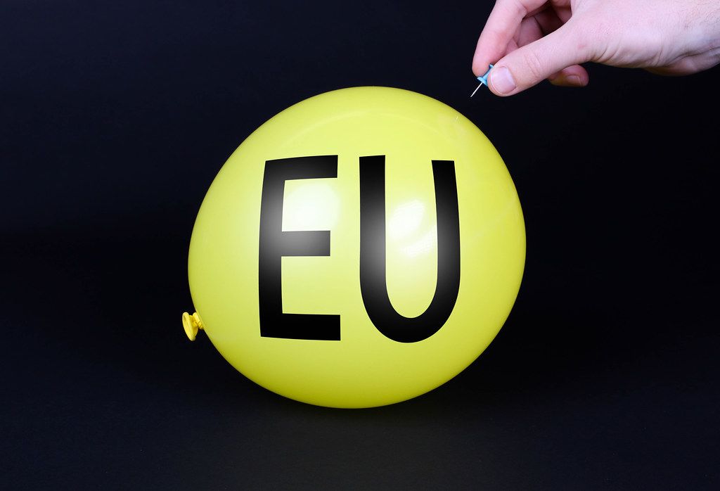 Hand uses a needle to burst a yellow balloon with EU text