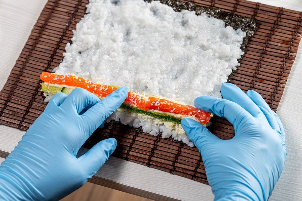 Hands in blue gloves spread the ingredients on a leaf of seaweed nori. Preparing sushi (Flip 2019)