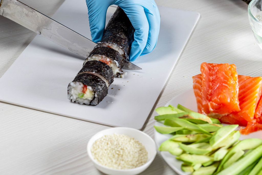 Hands in disposable gloves cutting sushi rolls on a cooking board