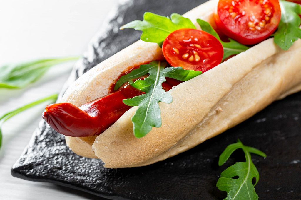Hot dog with smoke sausage, arugula leaves, ketchup and tomatoes on a black stone tray