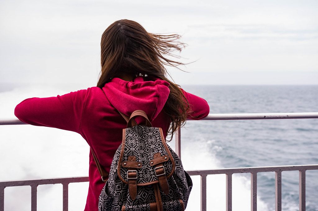 Lady in red pullover with backpack on her back looking out into the sea while the wind blows her hair (Flip 2019)