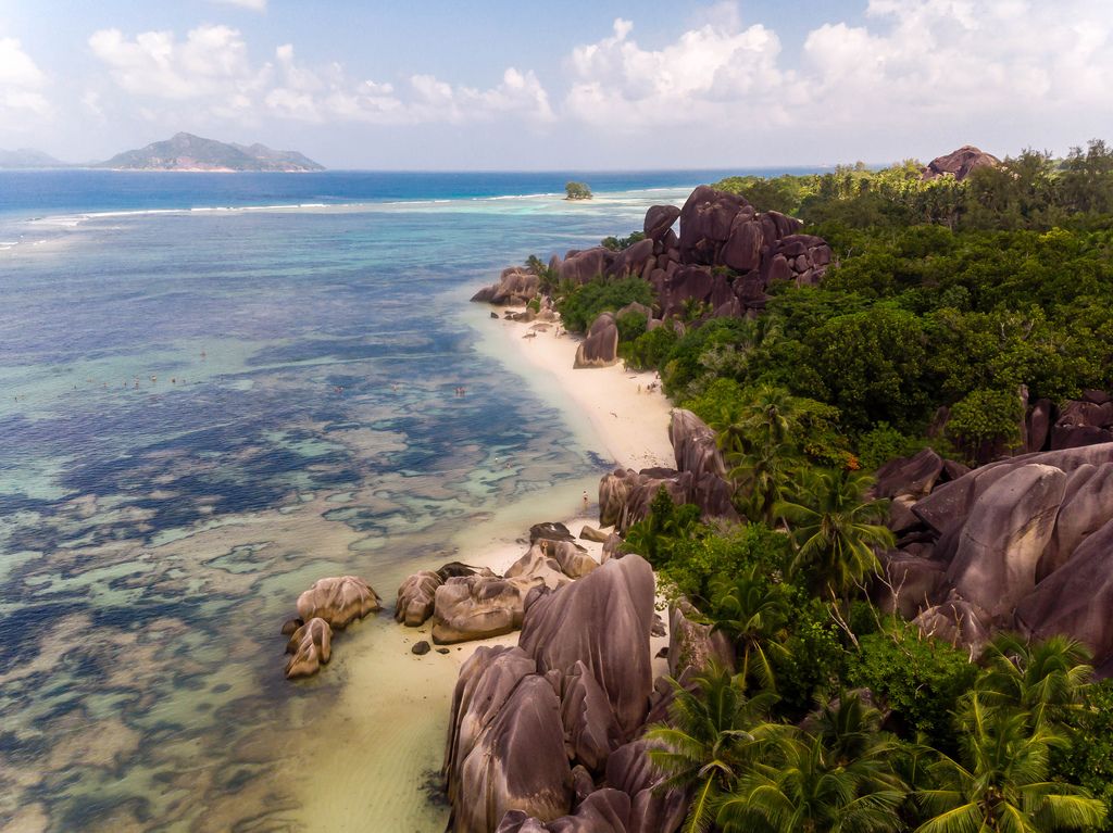 Luftbild zeigt Palmen, Granitfelsen und die Küste von La Digue und den Strand Anse Source d'Argent auf der Seychellen, mit badenden Menschen im Indischen Ozean