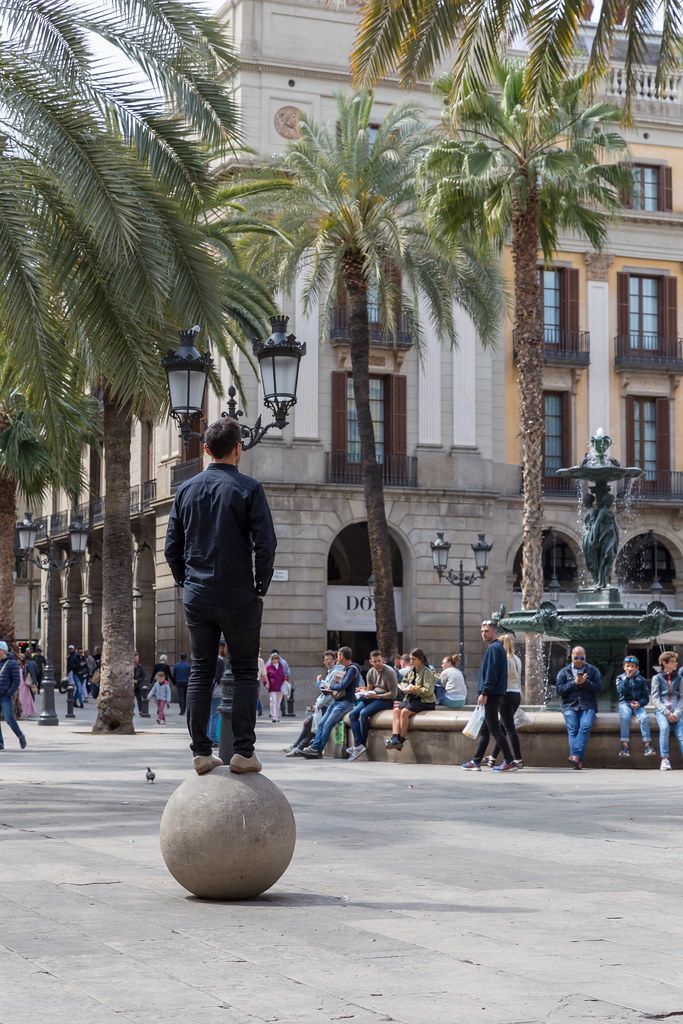Man balancing on a stone ball under palm trees, next to  fountain of the Three Graces and helmet lamps designed by Antoni Gaudi in Barcelona, Spain