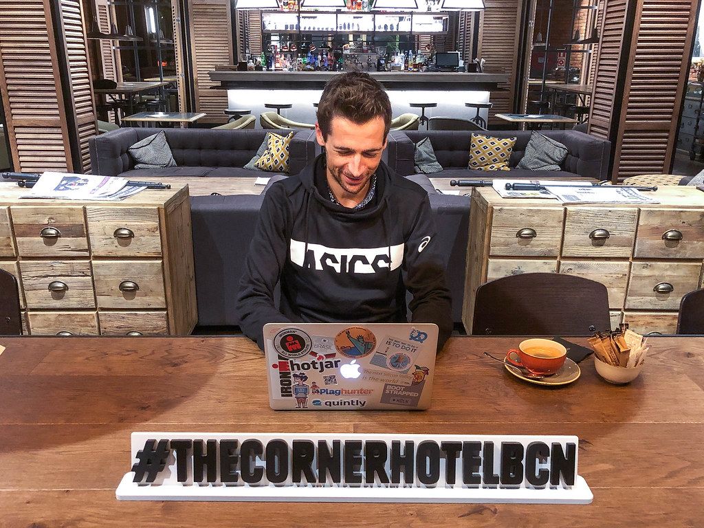 Man working on his Macbook with stickers, next to a coffee in the Corner Hotel Lounge, behind a #TheCornerHotelBCN Hashtag banner