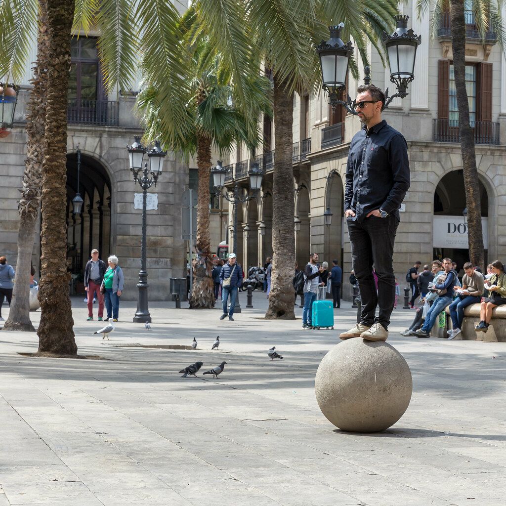 Mann balanciert auf steinerner Kugel vor den behelmten Laternen von Antoni Gaudí am historischen Placa Reial in Barcelona, Spanien