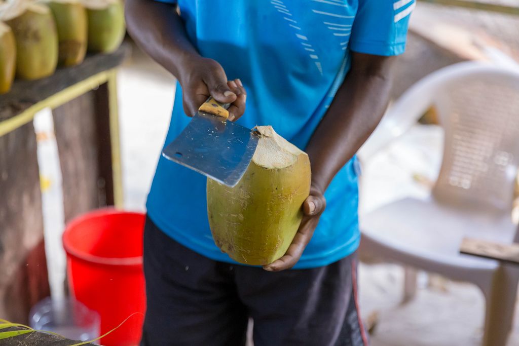 Mann mit Hackbeil beim Aufschneiden einer grünen Kokosnuss in einer Strandbar am Anse Source d'Argent Strand auf La Digue, Seychelleninsel