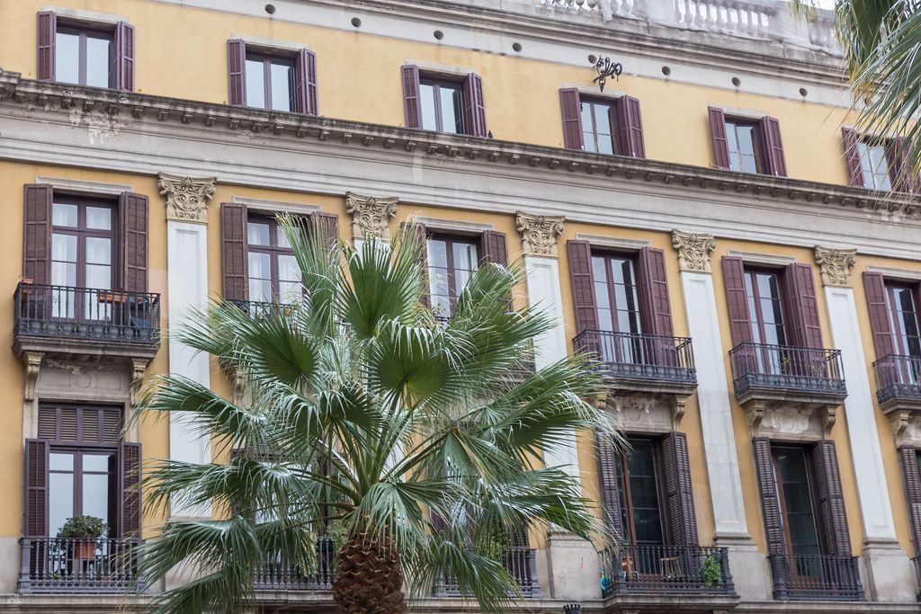 Mediterrane Architektur im klassizistischen Baustil mit Fensterläden aus Holz hinter Palmenblätter am Plaza Reial in Barcelona, Spanien
