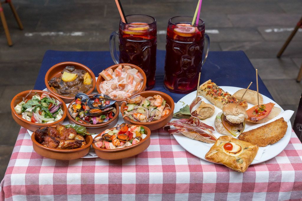 Mediterrane Delikatessen mit Muscheln, Shrimps, Gemüse und spanisches Brot mit Schinken vor roten Drinks auf einem kleinen Tisch in Barcelona