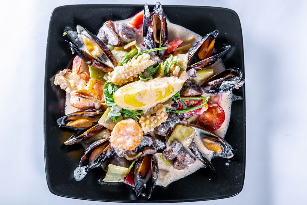Meeresfrüchte wie Muscheln Tintenfisch angerichtet mit Soße auf schwarzem Teller vor weißem Hintergrund