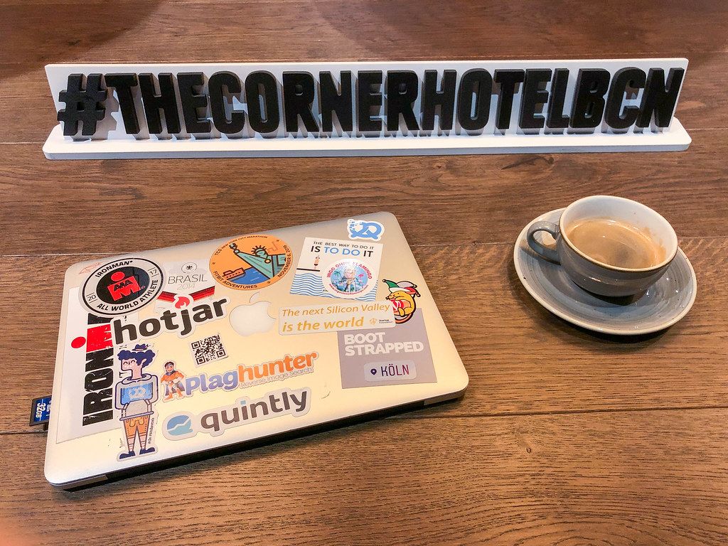 Mit Stickern beklebtes Macbook von Apple neben einer Tasse Kaffee vor dem Hashtag-Schild #TheCornerHotelBCN in Barcelona, Spanien