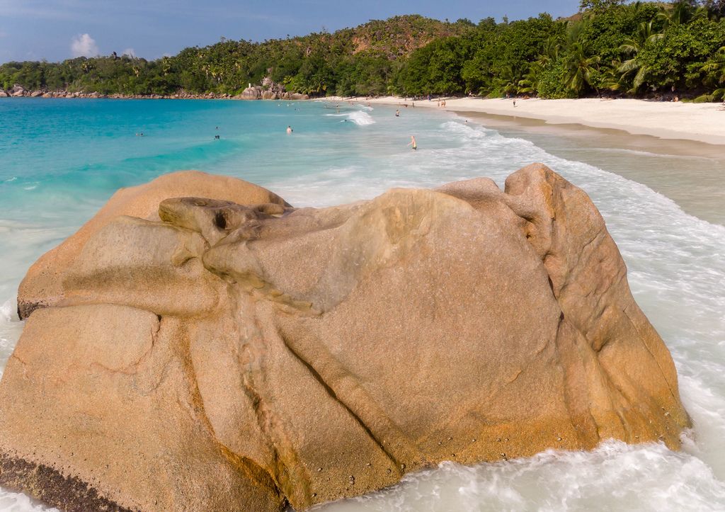 Nahaufnahme eines braunen Granitfelsen am Strand Anse Lazio auf Praslin, Seychellen, mit badenden Touristen im Hintergrund