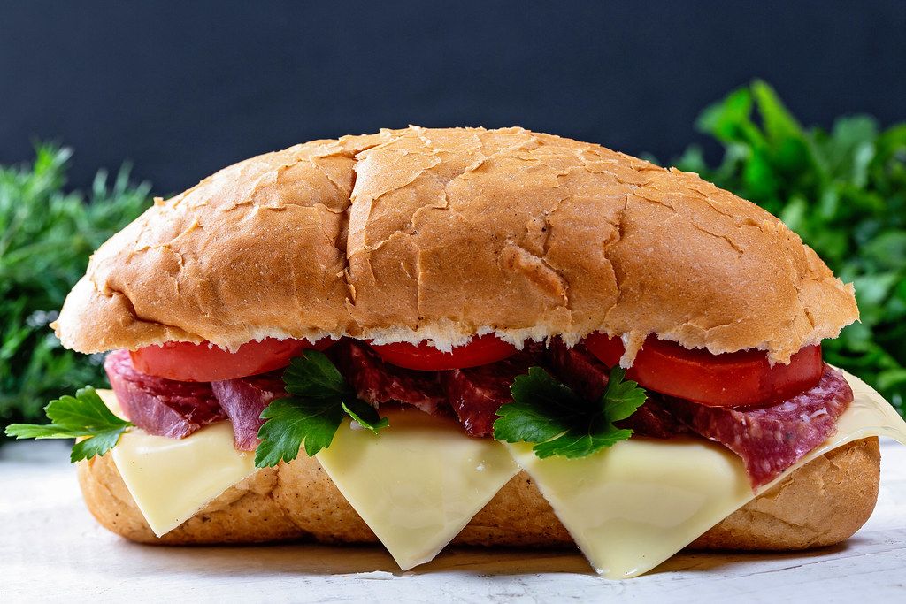 Nahaufnahme von einem Sandwich-Baguette mit Salami, Tomatenscheiben, Käse und grünen Kräutern
