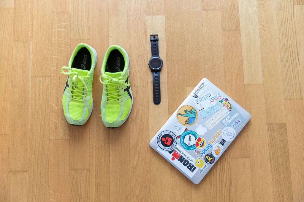 Neongelbe Asics Laufschuhe mit Garmin Armbanduhr und Macbook auf Parkettboden