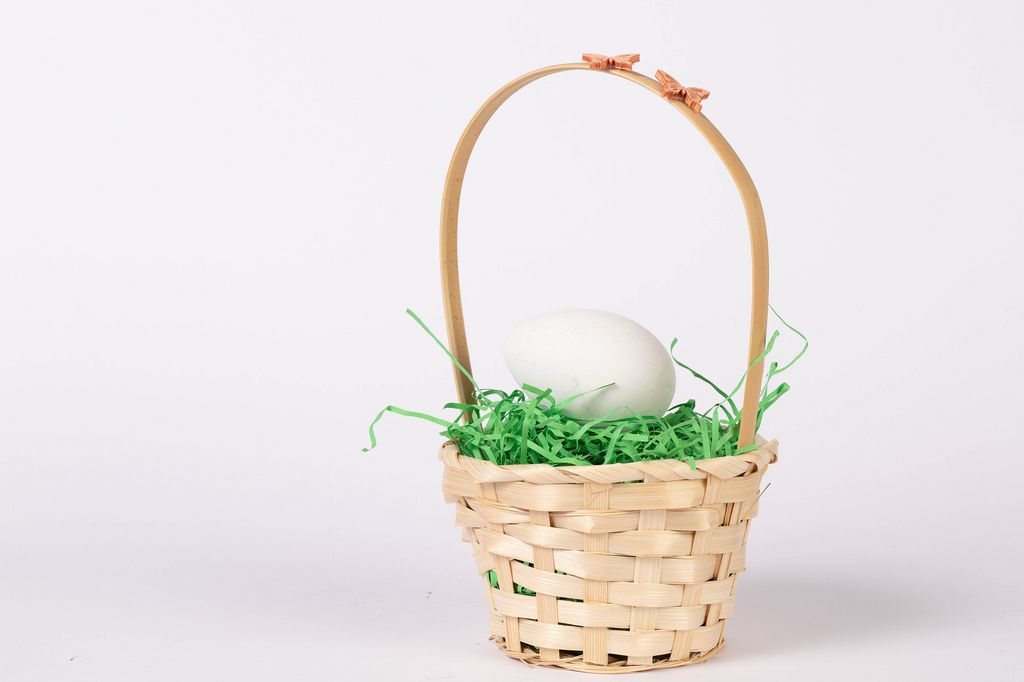 Osterdekoration Bastkörbchen mit Henkel gefüllt mit Ostergras und unbemaltem Ei vor weißem Hintergrund