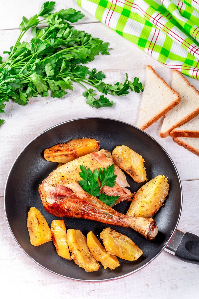 Pan with fried chicken leg and potatoes on a white wooden table with bread and herbs