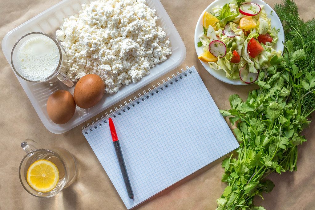 Planning diet concept. Flat lay view of notepad with a pen and organic healthy food: cottage cheese, eggs, greens, salad, apple, and greens on brown paper (Flip 2019)