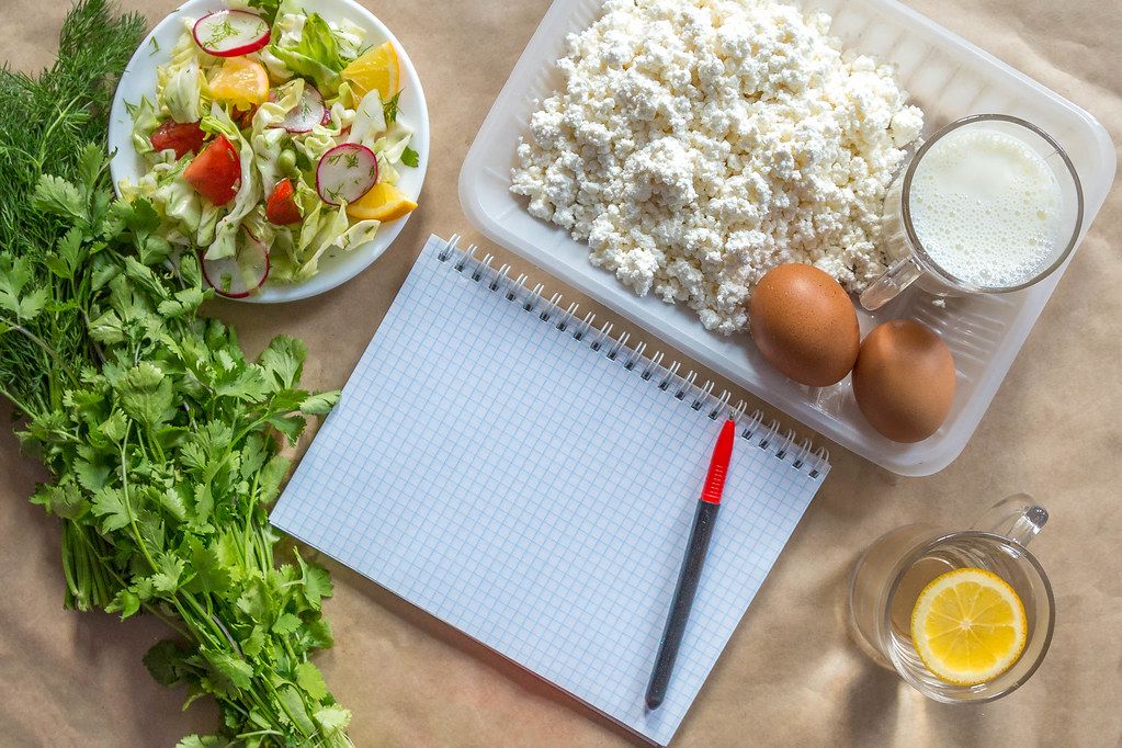 Planning diet concept. Flat lay view of notepad with a pen and organic healthy food: cottage cheese, eggs, greens, salad, apple, and greens on brown paper
